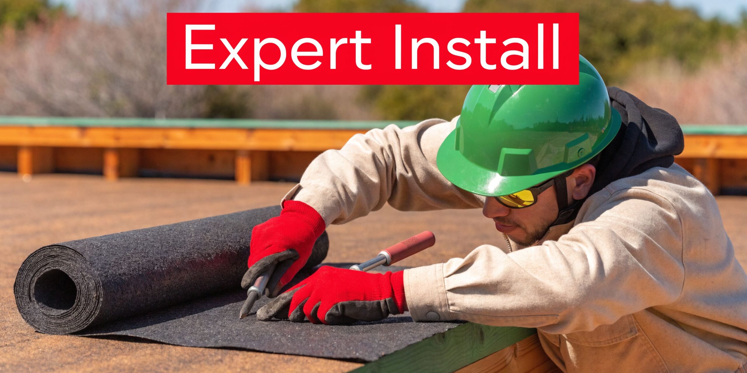 A professional construction worker wearing a green hard hat installs roll roofing on a flat roof.